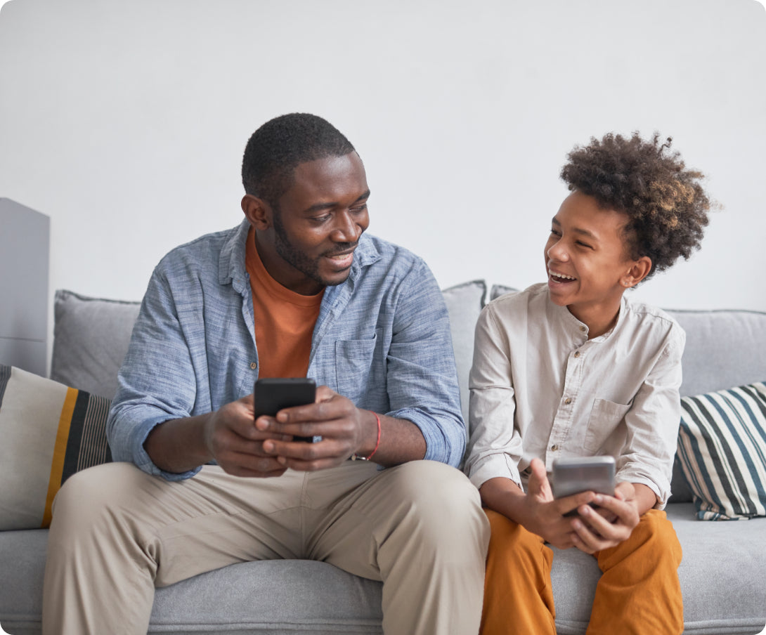 A man on the couch with his son, both are on their phones
